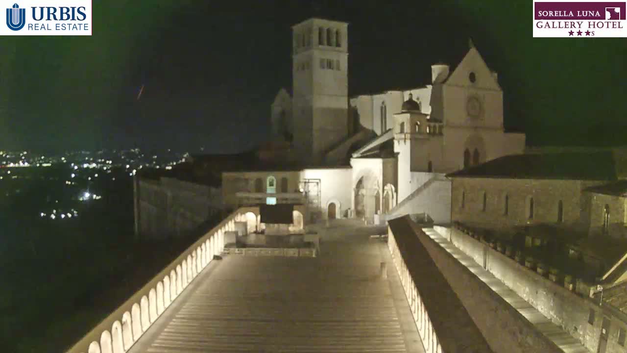 A large, illuminated stone structure, possibly a church or basilica, stands at night against a dark sky, overlooking a city with distant lights.