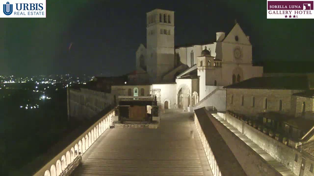 A large, illuminated stone building complex stands at night, overlooking a city with many lights below.