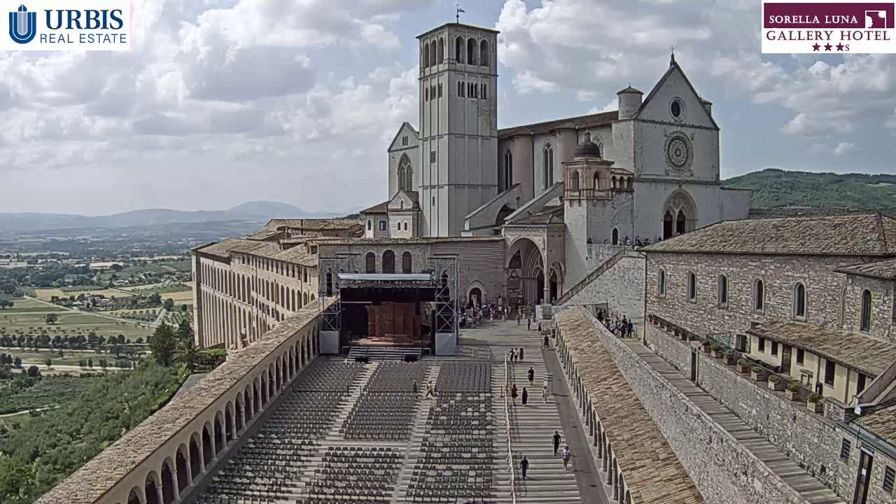 A large stone basilica with an outdoor amphitheater filled with chairs sits on a hill overlooking a sprawling green valley under a partly cloudy sky.
