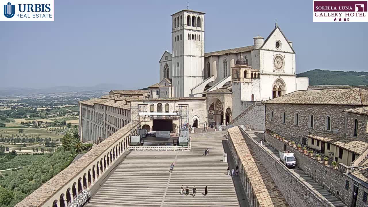 A large, light-colored basilica and adjacent buildings stand on a hill overlooking a sprawling landscape under a clear, sunny sky.
