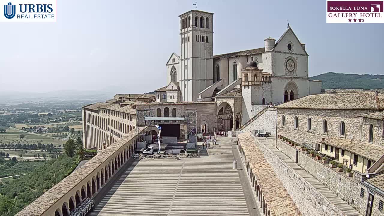 A large stone basilica and adjoining buildings are situated on a hill overlooking a valley under a clear, sunny sky, with a paved plaza in front featuring a stage setup.