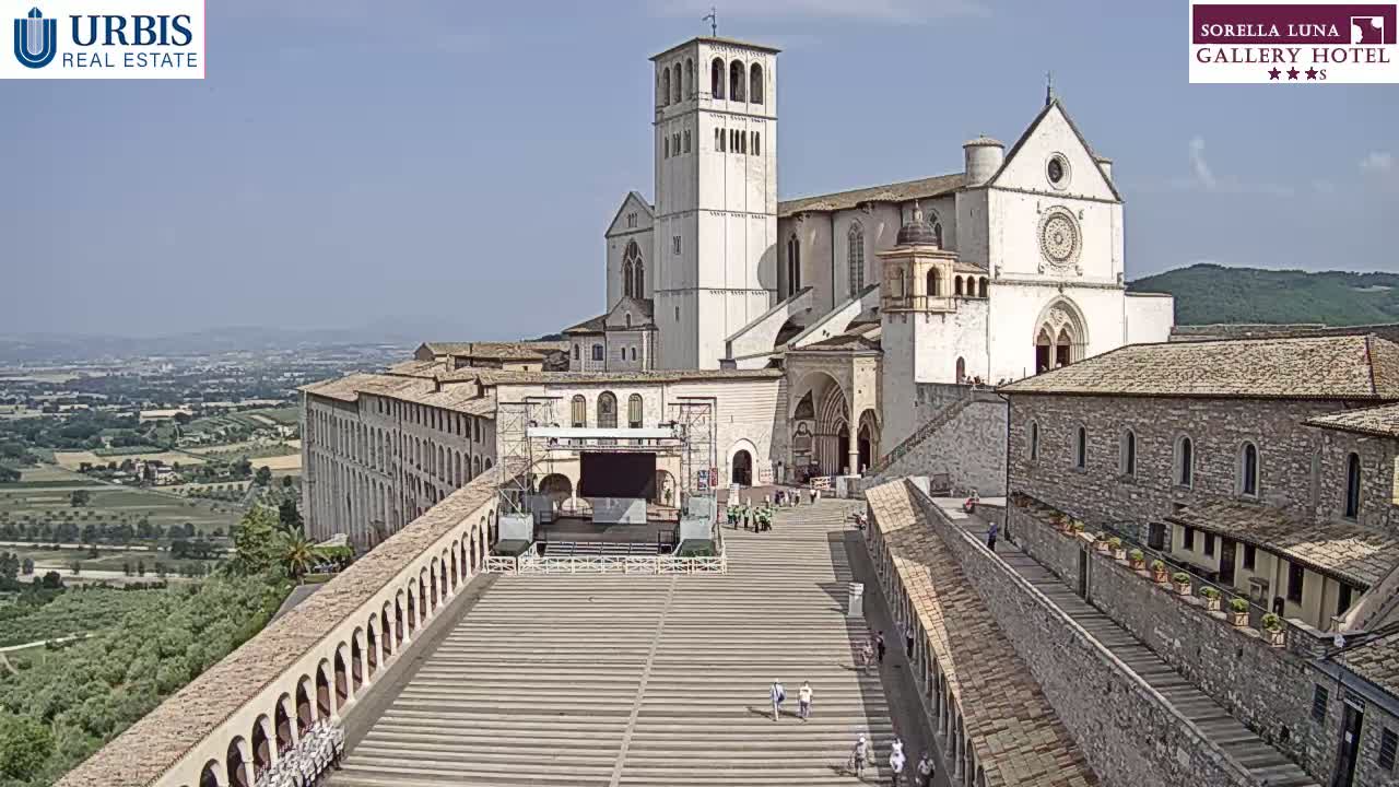 A large, light-colored basilica stands on a hill overlooking a sprawling landscape under a clear, sunny sky.