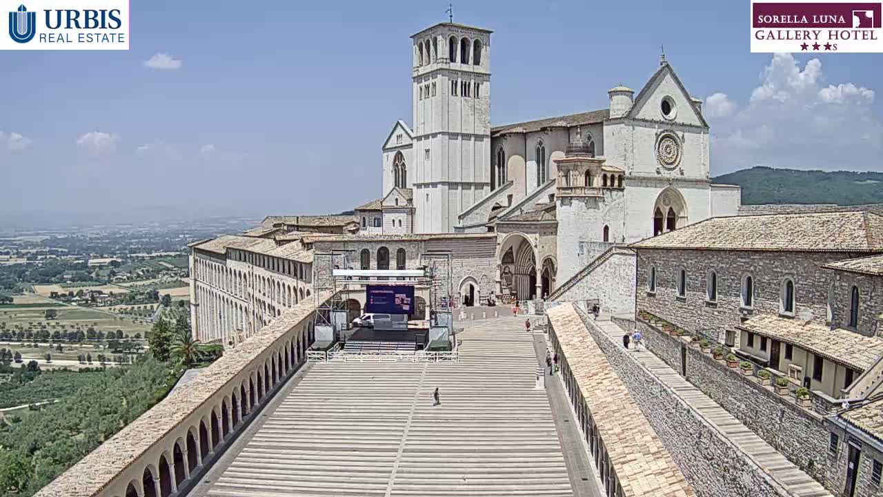 A large, light-colored stone basilica sits atop a hill on a sunny day, with a wide plaza and walkways leading to it, overlooking a verdant valley.