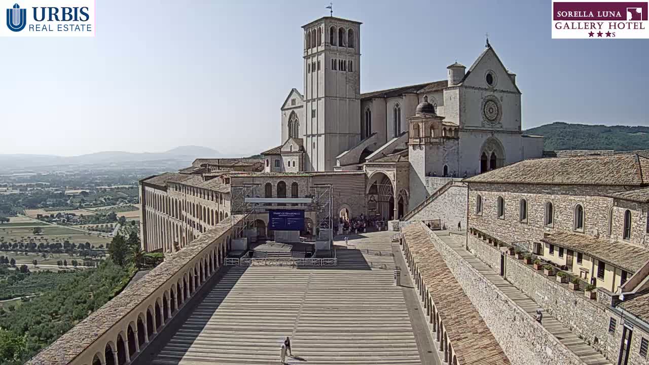 A large, light-colored basilica with a tall bell tower stands prominently in a sunlit plaza, flanked by long, arched walkways, overlooking a sprawling landscape under a clear sky.