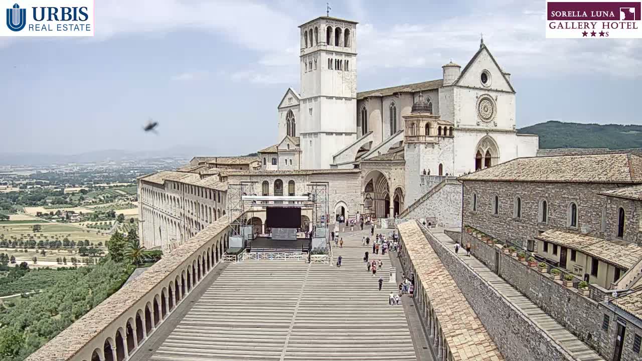 A large, white basilica overlooks a paved plaza with several people walking on a sunny day, surrounded by older stone buildings and a verdant landscape in the distance.