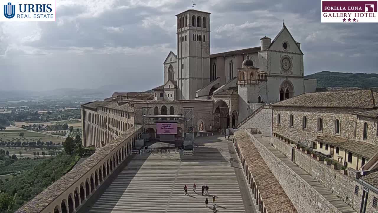 A large stone basilica and adjoining buildings are seen from above on a partly cloudy day, with a wide paved plaza in front and a view of rolling hills in the distance.