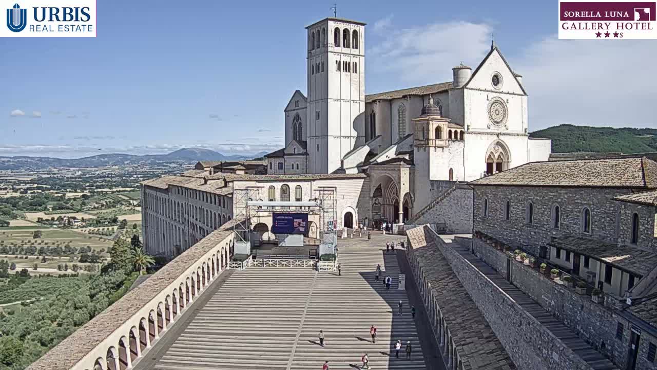 A large, light-colored basilica and adjacent buildings are situated on a hilltop overlooking a verdant valley under a clear, sunny sky.