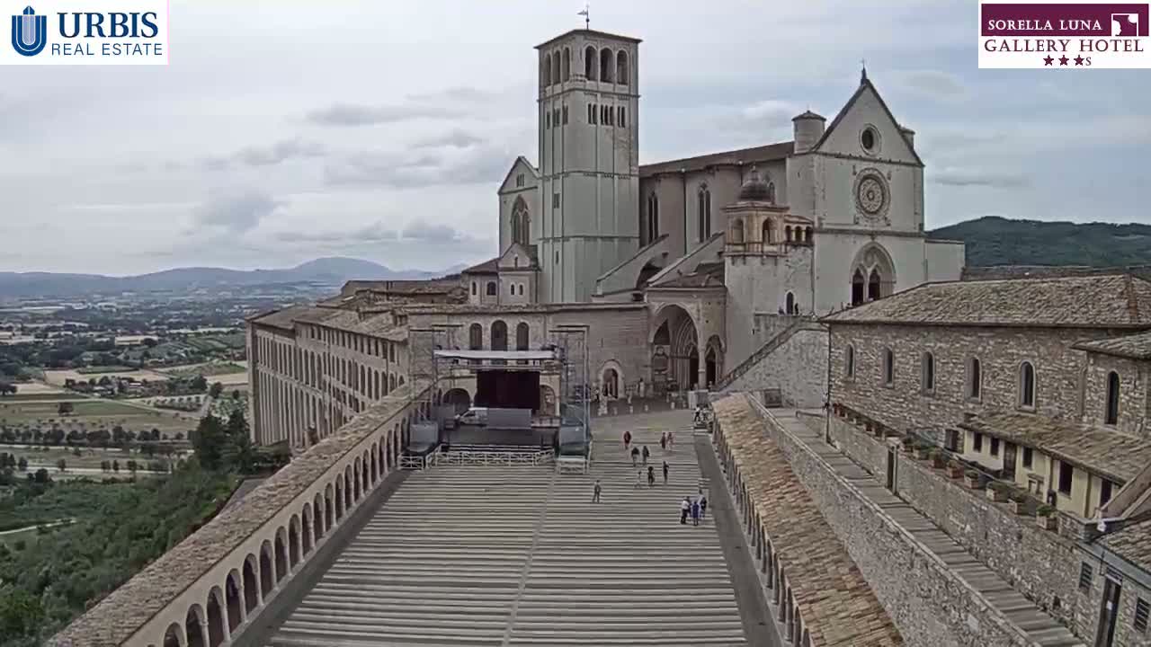 A large stone basilica sits atop a hill overlooking a valley under an overcast sky, with a stage and seating area in the foreground.