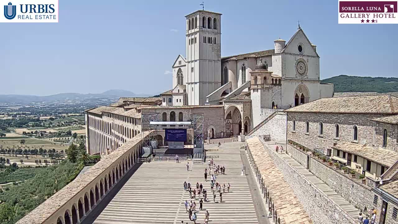 A large stone basilica and surrounding buildings are situated on a hill overlooking a valley on a sunny day, with a few people visible in the plaza below.