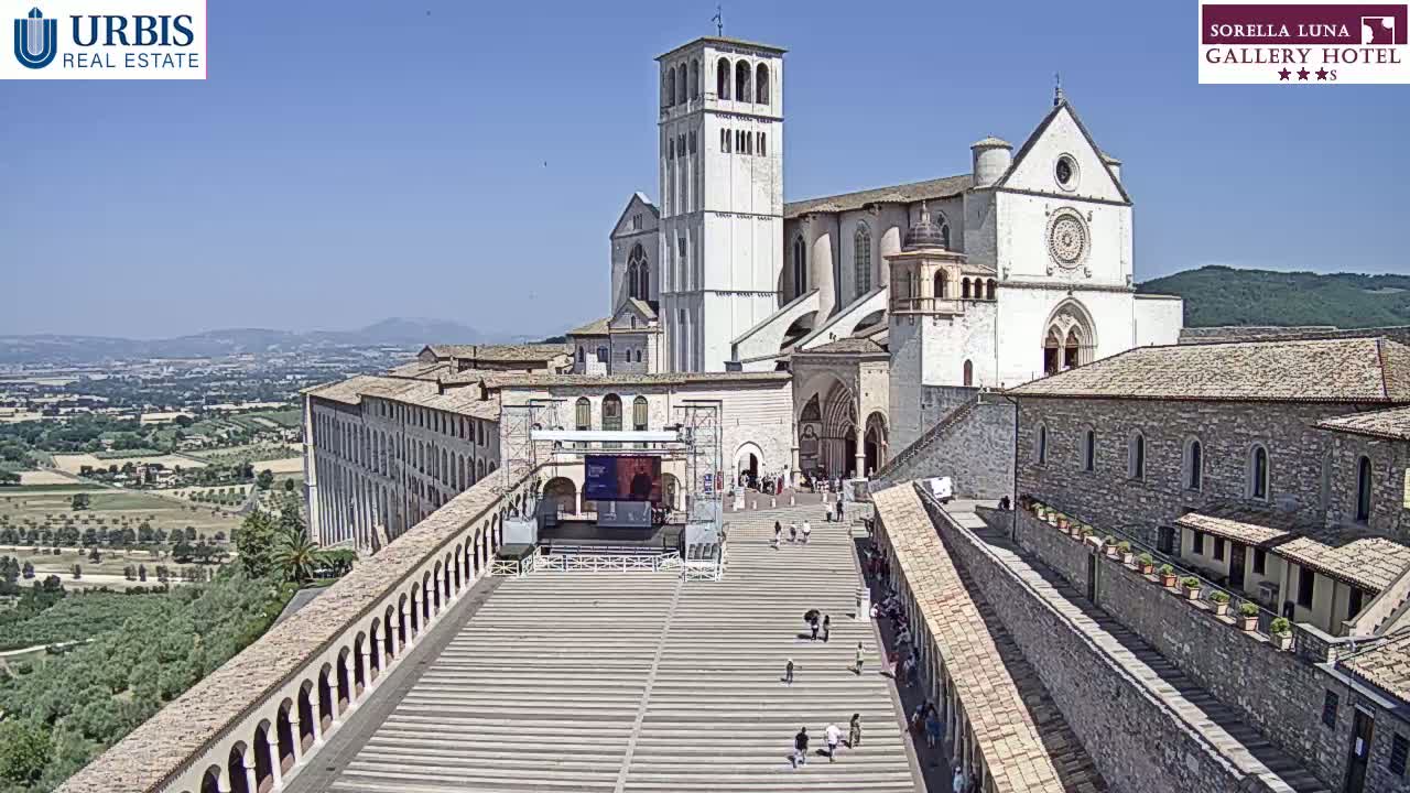 A large stone basilica and adjoining buildings are shown on a sunny day, with a plaza in front containing a few people and a large stage.