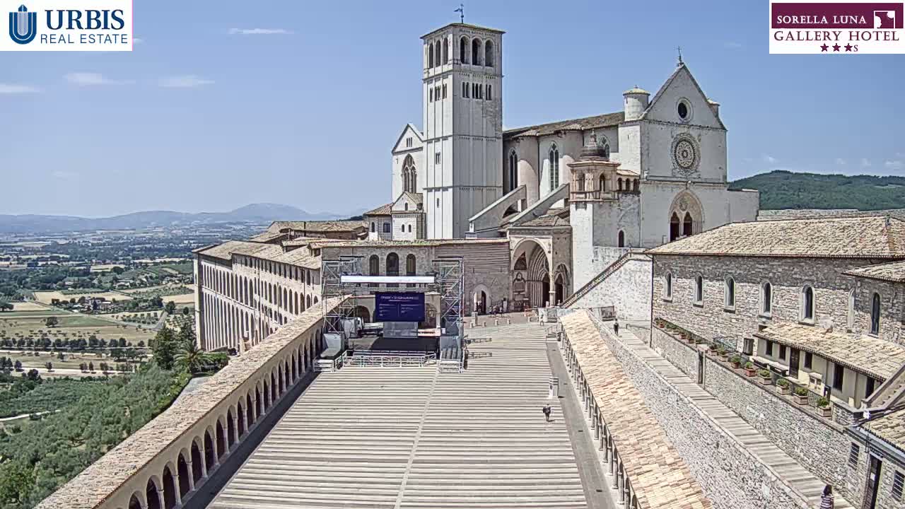 A large stone basilica and surrounding buildings are viewed from above on a sunny day, overlooking a paved plaza and a distant landscape.