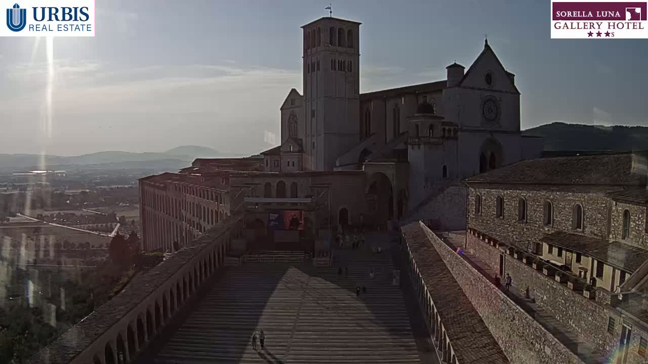 A large stone basilica and adjoining buildings are situated on a hill overlooking a town under a partly sunny sky.