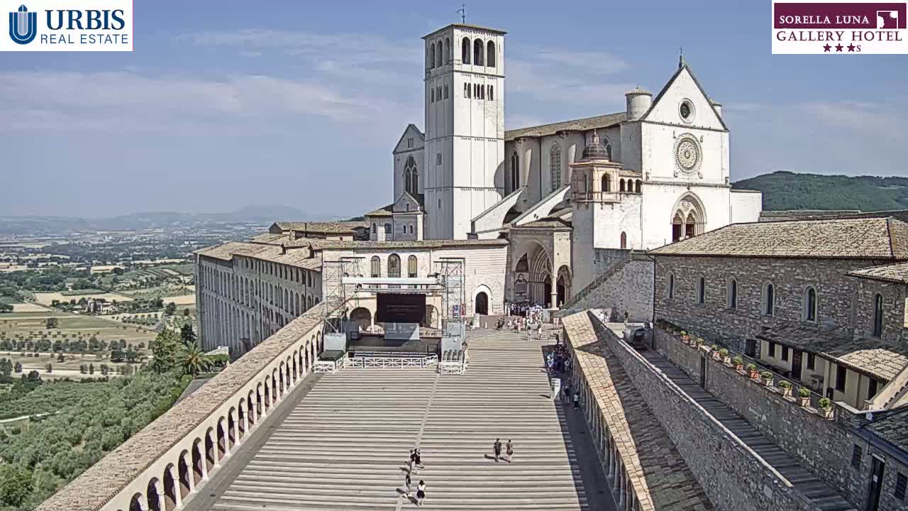 A large stone basilica and adjacent buildings are seen from above on a sunny day, with a wide staircase leading to the main entrance and a few people walking on it.