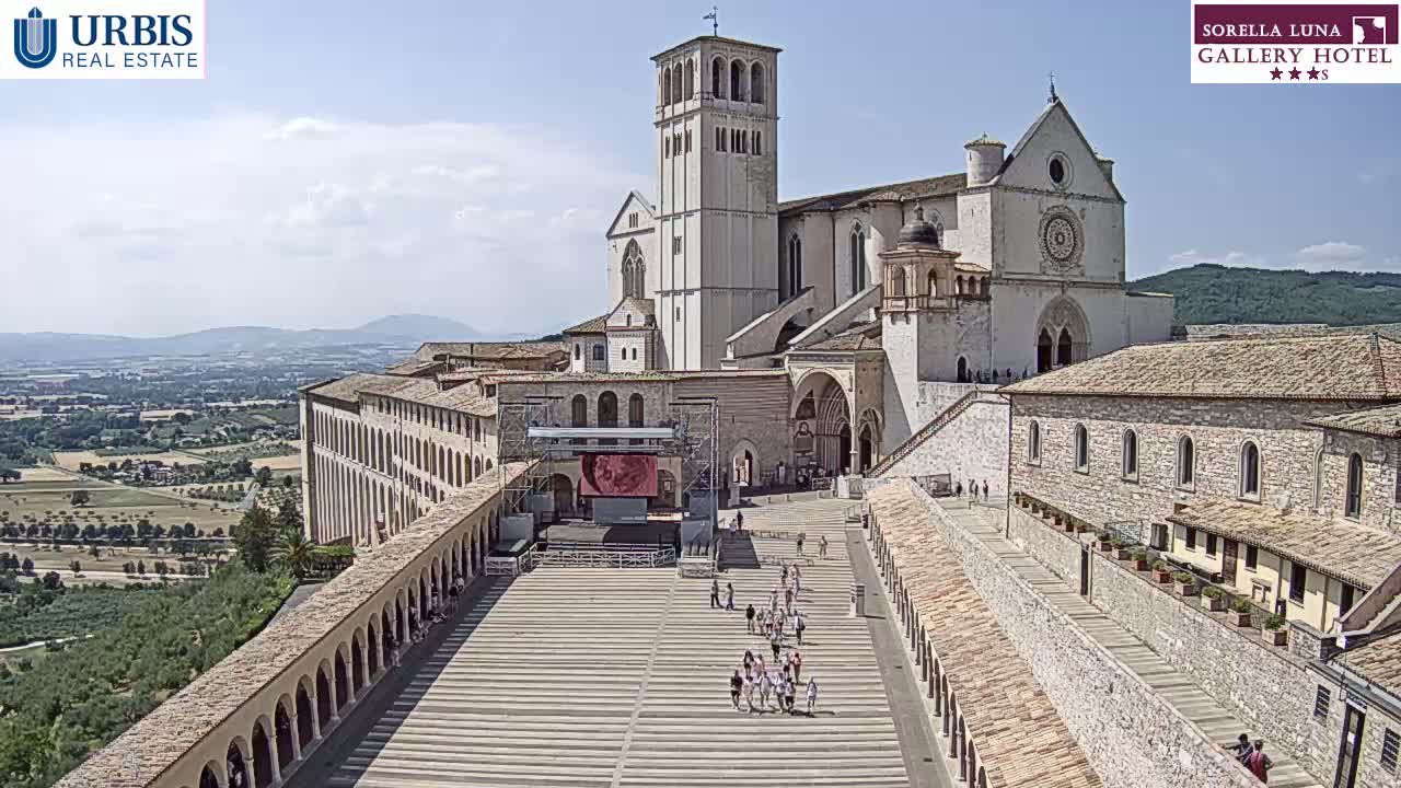 A large stone basilica and adjacent buildings are situated on a hilltop overlooking a valley under a clear, sunny sky, with a paved plaza containing a few people in the foreground.