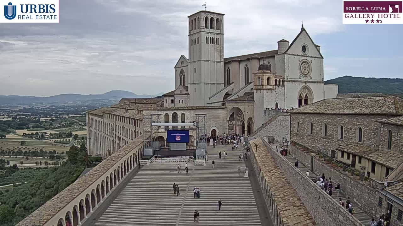 A large stone basilica and surrounding buildings are viewed from above on an overcast day, with many people walking on the expansive plaza and stairs leading to the church.