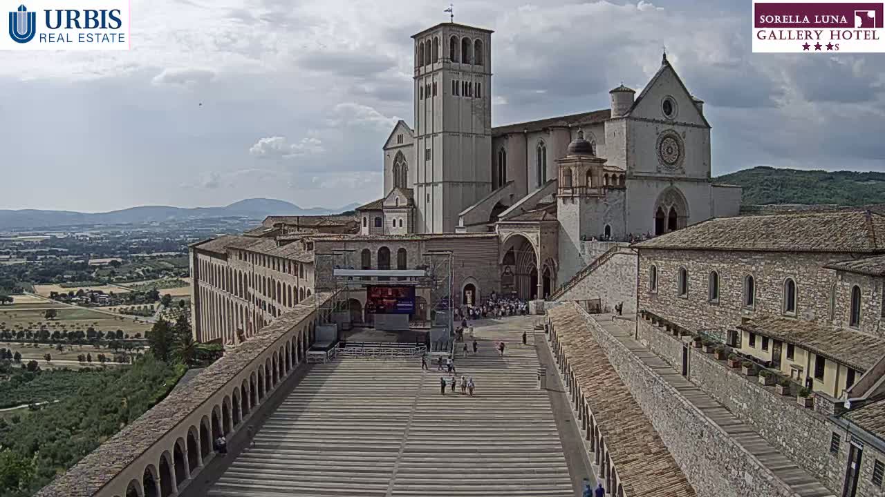A large stone basilica and adjoining buildings stand on a hill overlooking a verdant valley under a partly cloudy sky.