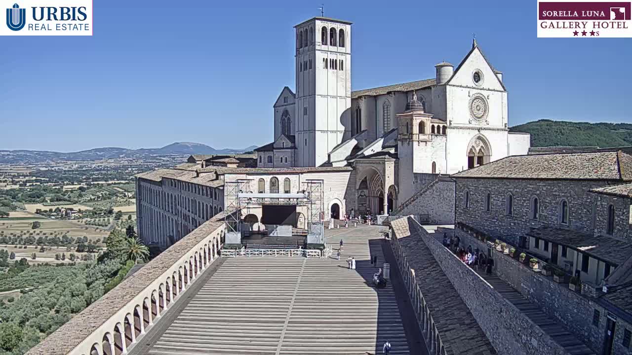 A large, light-colored basilica sits atop a hill on a sunny day, overlooking a sprawling landscape of green hills and valleys.