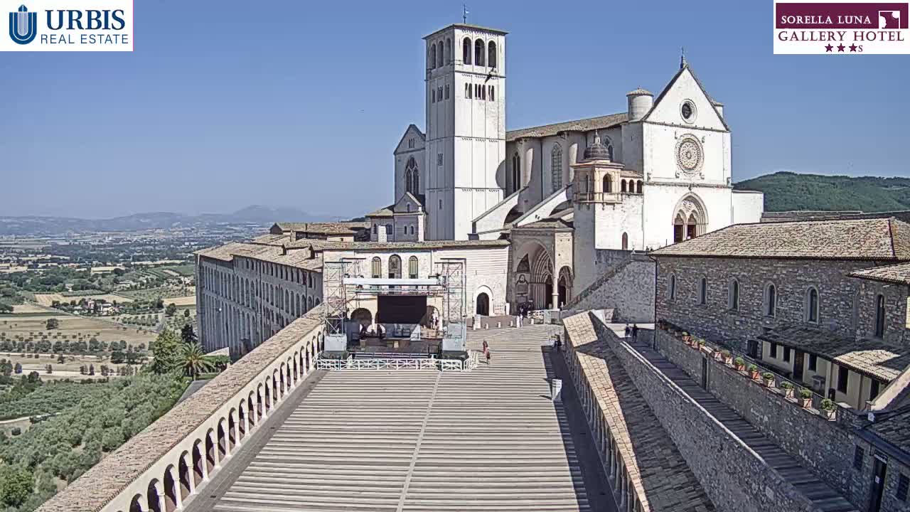A large stone basilica sits atop a hill on a sunny day, overlooking a vast landscape of olive groves and distant mountains.