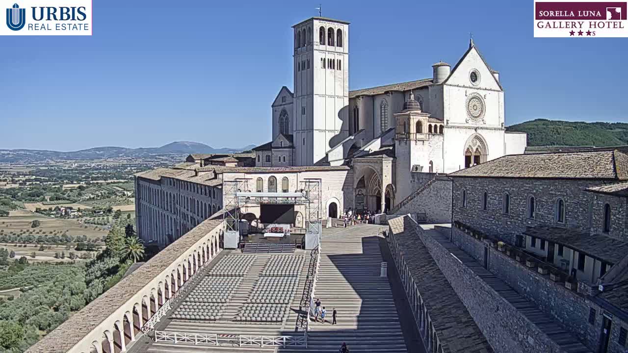 A large stone basilica sits atop a hill overlooking a sunlit valley, with a wide staircase leading down to a large outdoor seating area.