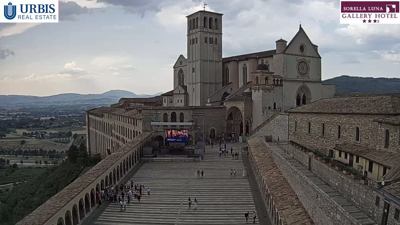 A large stone basilica sits atop a hill overlooking a sunlit valley, with a wide staircase leading down to a large outdoor seating area.