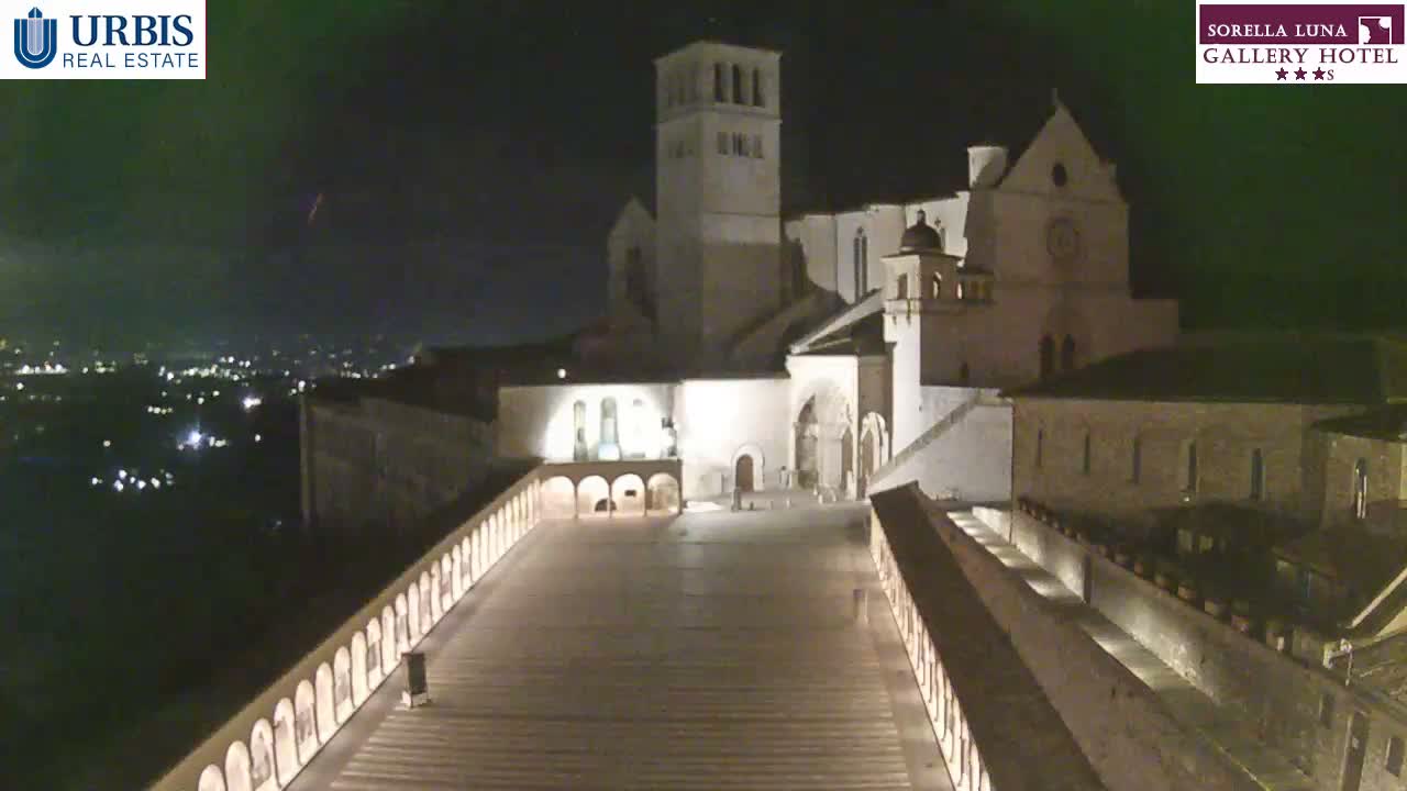 An illuminated grand basilica stands prominently above a lit paved square at night, with distant city lights twinkling under a clear sky.