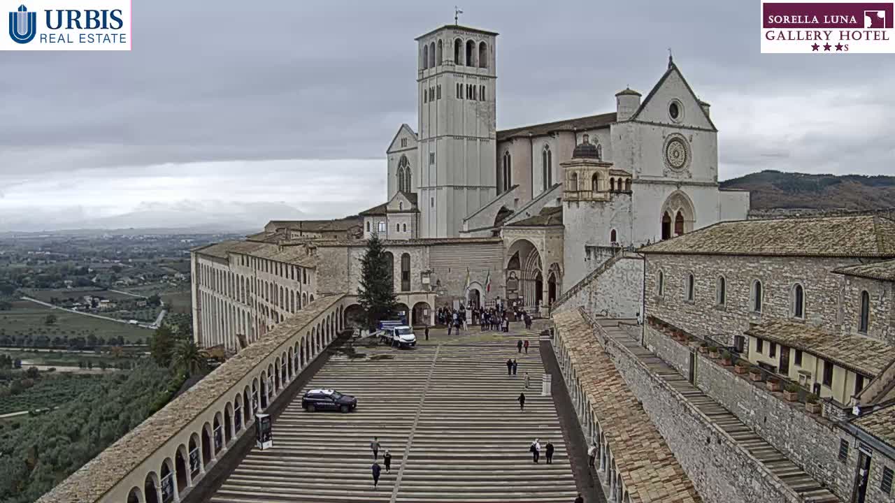 A grand, light-colored basilica with a tall bell tower stands prominently on a hillside, overlooking a valley under an overcast sky, with numerous people gathered on the wide tiered steps leading up to its entrance.