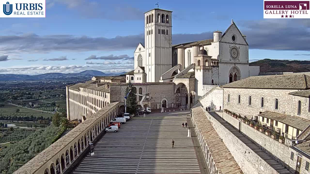 A grand, light-colored basilica with a tall bell tower stands prominently on a hillside, overlooking a valley under an overcast sky, with numerous people gathered on the wide tiered steps leading up to its entrance.