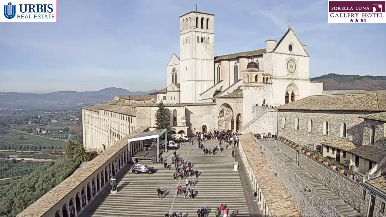 A historic basilica with a tall bell tower stands prominently above a wide, tiered plaza filled with people, all set against a backdrop of a sprawling valley and distant mountains under a clear, sunny sky.