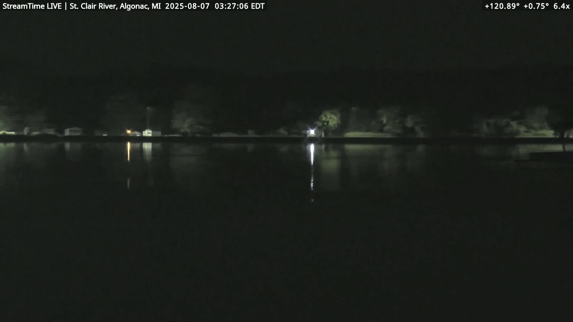 A dark nighttime view shows a calm body of water reflecting a dimly lit shoreline with trees and buildings.