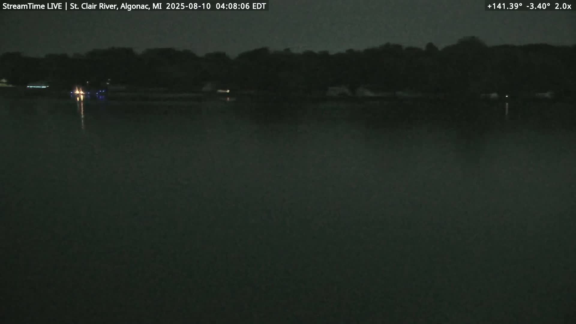 A dark, nighttime view of a calm body of water with a tree-lined shore in the background.