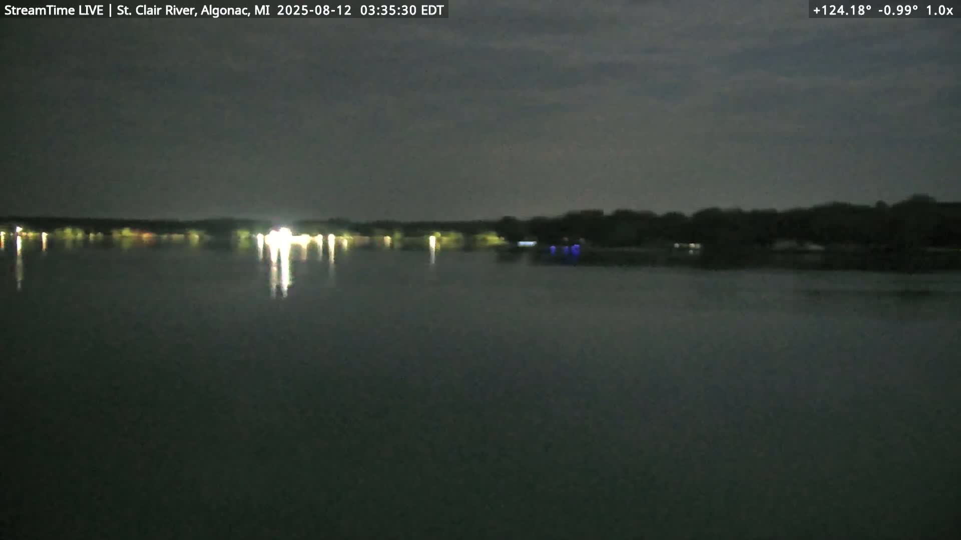 A dark, nighttime view of a calm body of water reflecting the lights of a distant shoreline under a slightly cloudy sky.