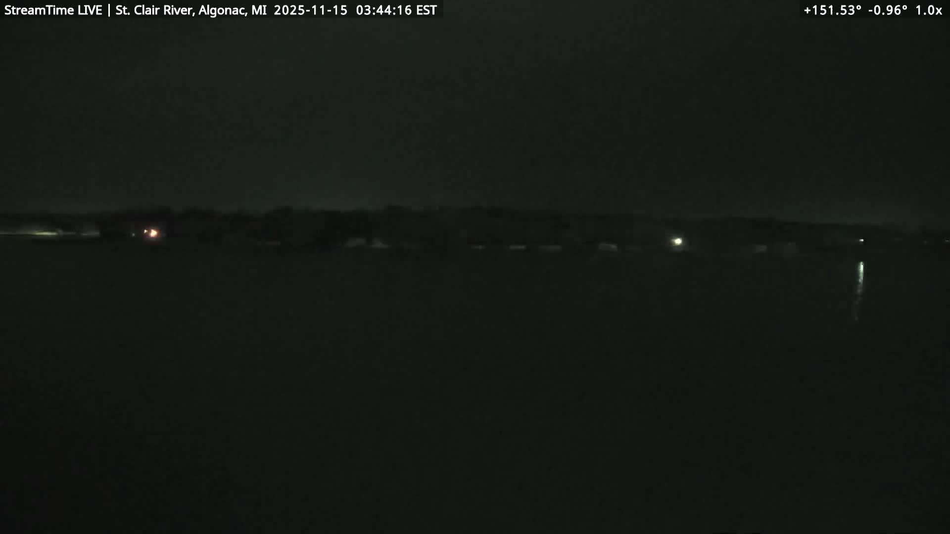 A dark, clear night scene captures a body of water with scattered lights visible on the distant shore and a distinct light reflection stretching across the water on the right.