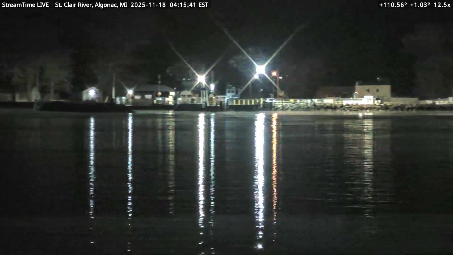 A dark, clear night scene captures a body of water with scattered lights visible on the distant shore and a distinct light reflection stretching across the water on the right.