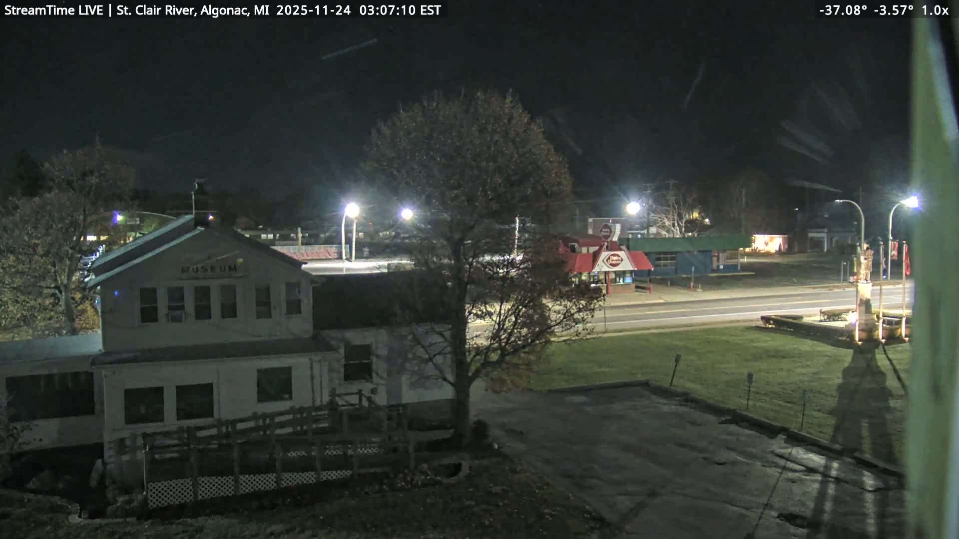 A nighttime scene features a multi-story white building and a large, mostly bare tree on the left, with a brightly lit road and other commercial buildings visible across it on the right, all under a clear, dark sky.