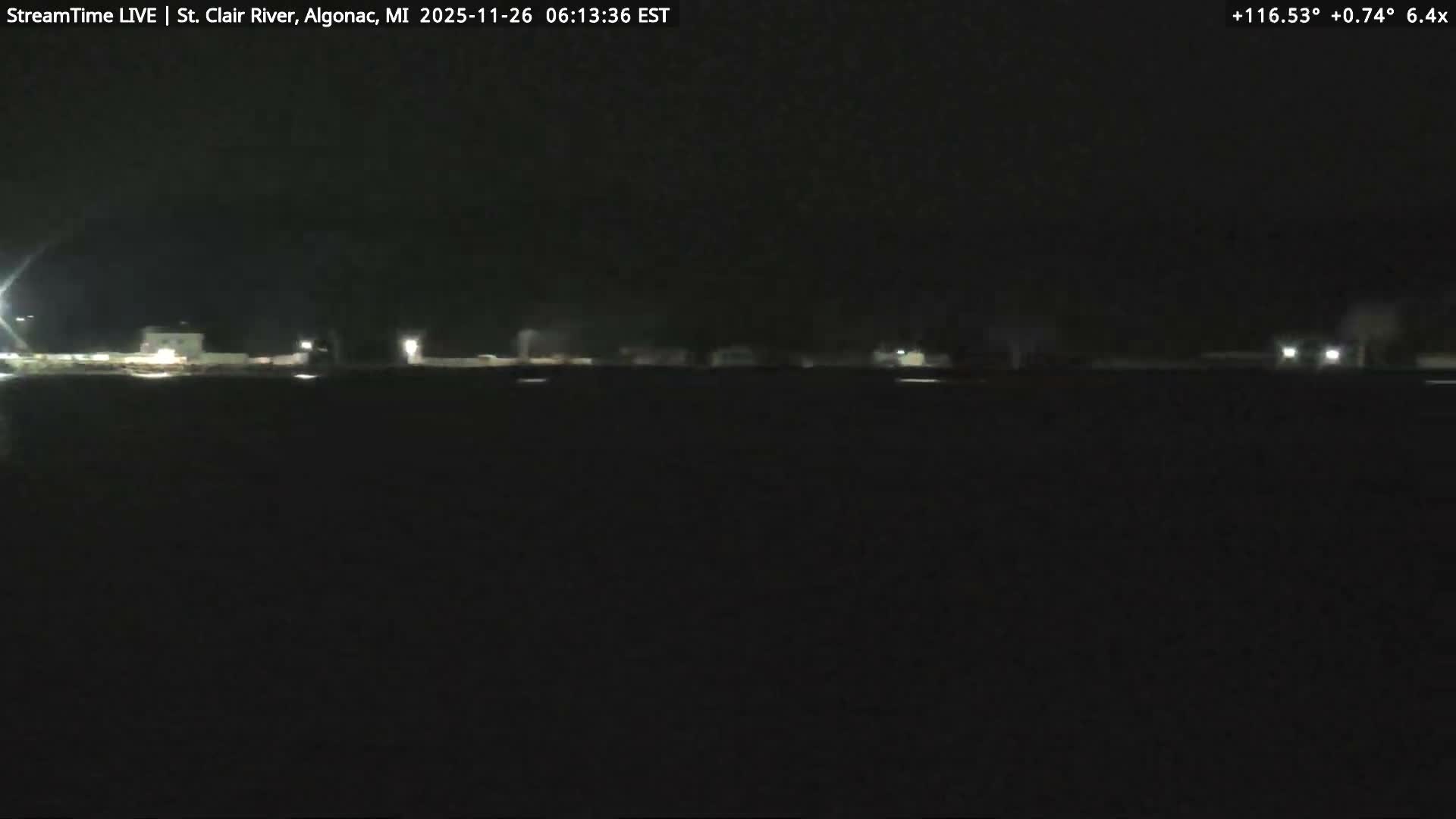 A nighttime scene features a multi-story white building and a large, mostly bare tree on the left, with a brightly lit road and other commercial buildings visible across it on the right, all under a clear, dark sky.