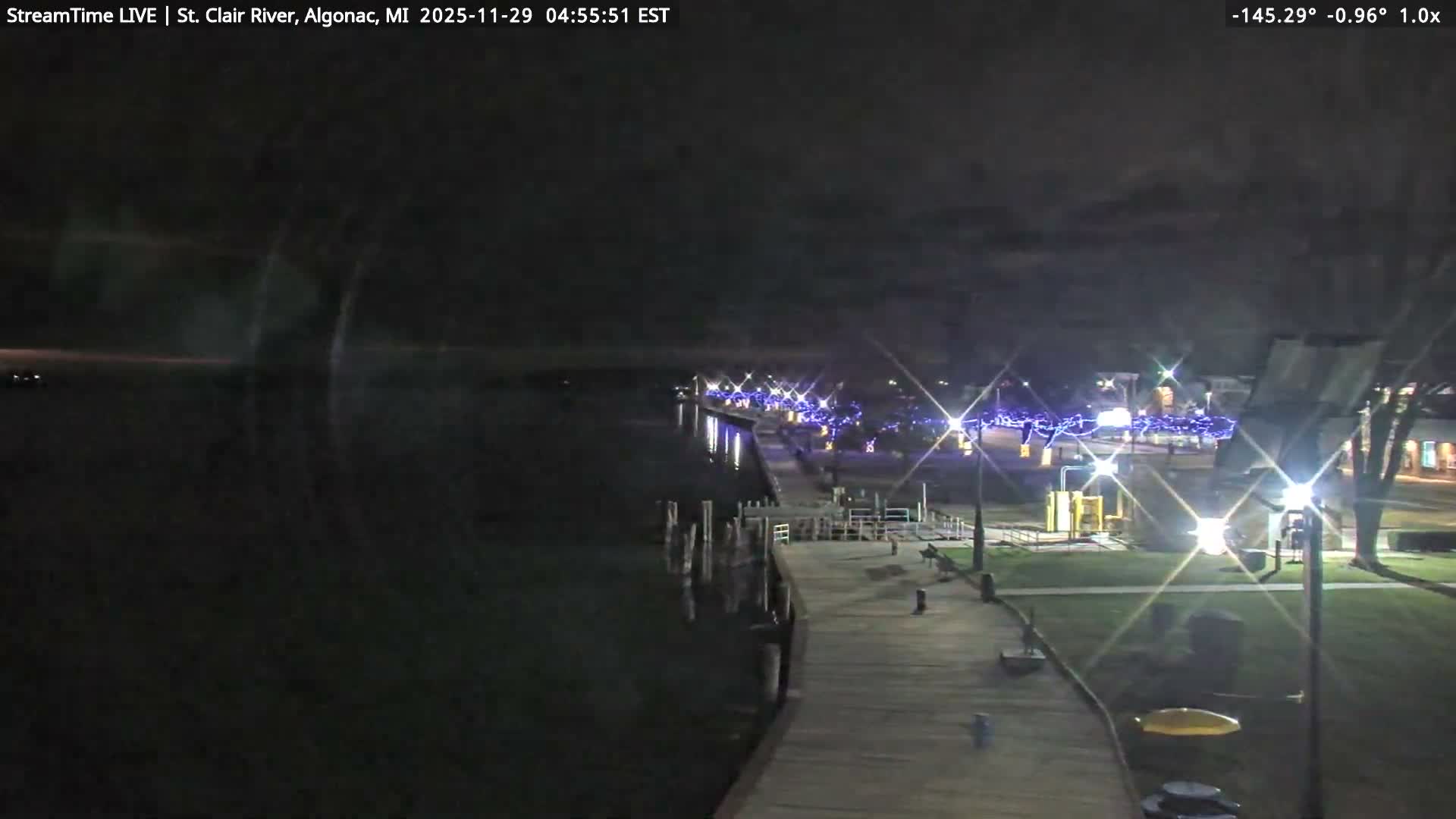 An outdoor nighttime scene reveals a well-lit wooden pier bordering a dark body of water, with festive blue string lights adorning trees and structures ashore under a clear, dark sky.