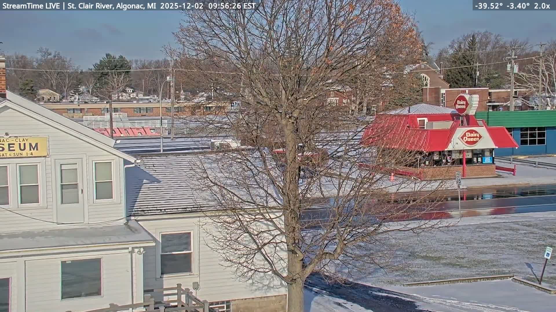 A cold, sunny winter day shows a snow-dusted landscape featuring a large bare tree dominating the center, a white two-story building on the left, and a red-roofed eatery on the right, with other structures and bare trees in the background under a clear blue sky.