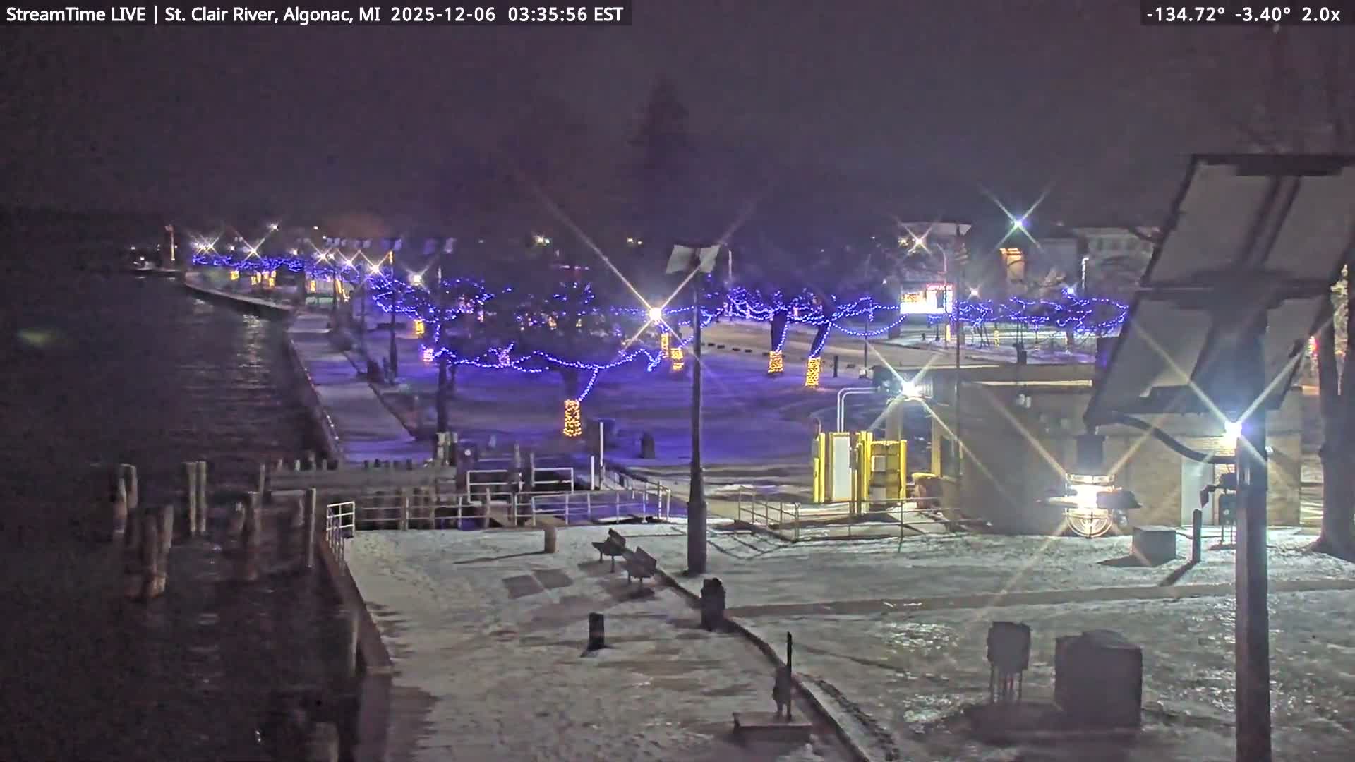 A snow-dusted riverfront promenade is brightly illuminated at night by streetlights and trees adorned with blue and orange festive lights under a clear sky.