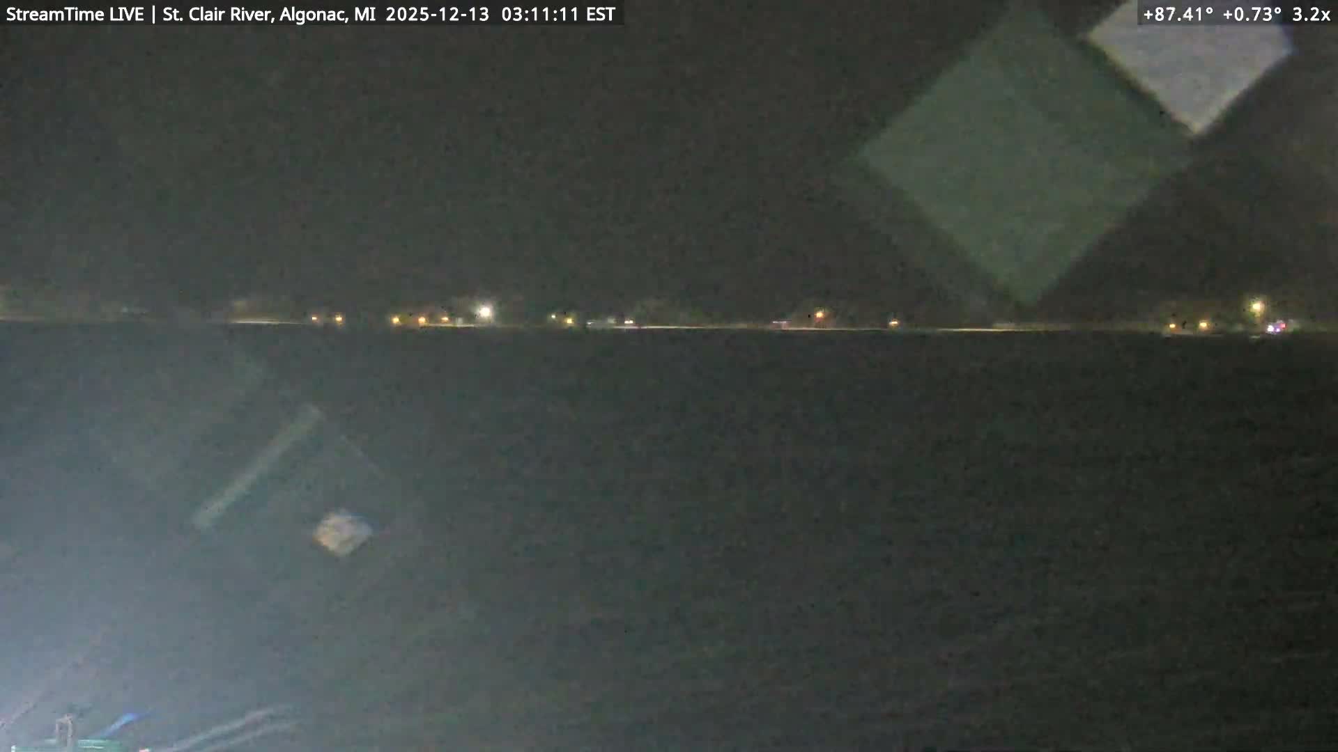 A dark nighttime view shows a body of water in the foreground with a line of distant lights visible across the opposite shore under clear conditions.