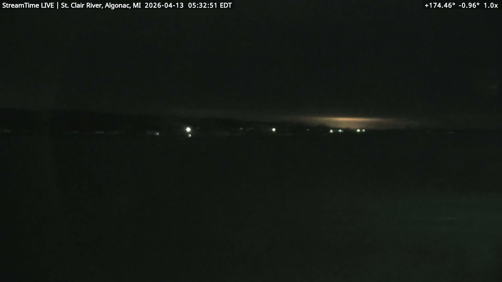 A dark nighttime view shows a body of water in the foreground with a line of distant lights visible across the opposite shore under clear conditions.