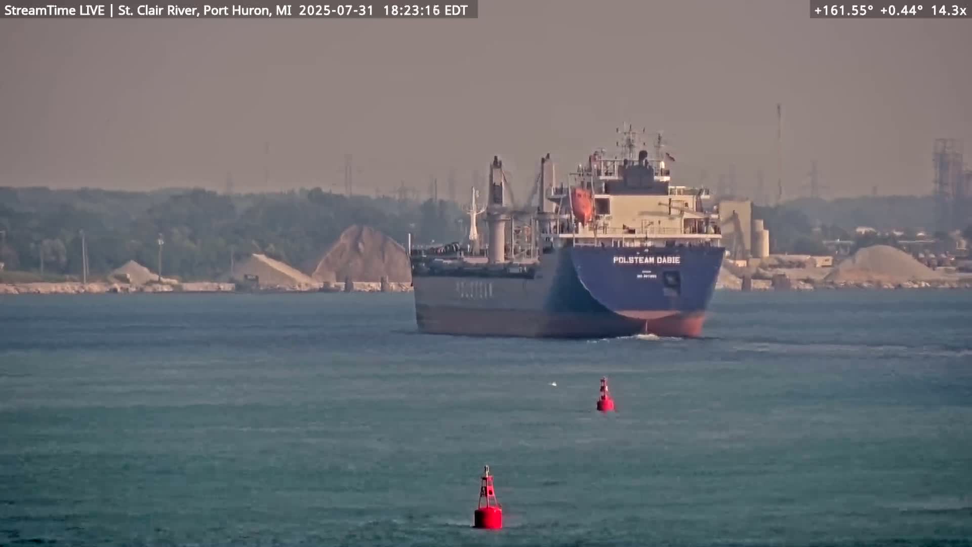 A large cargo ship sails on calm, hazy water past two red navigation buoys, with a shoreline visible in the background.