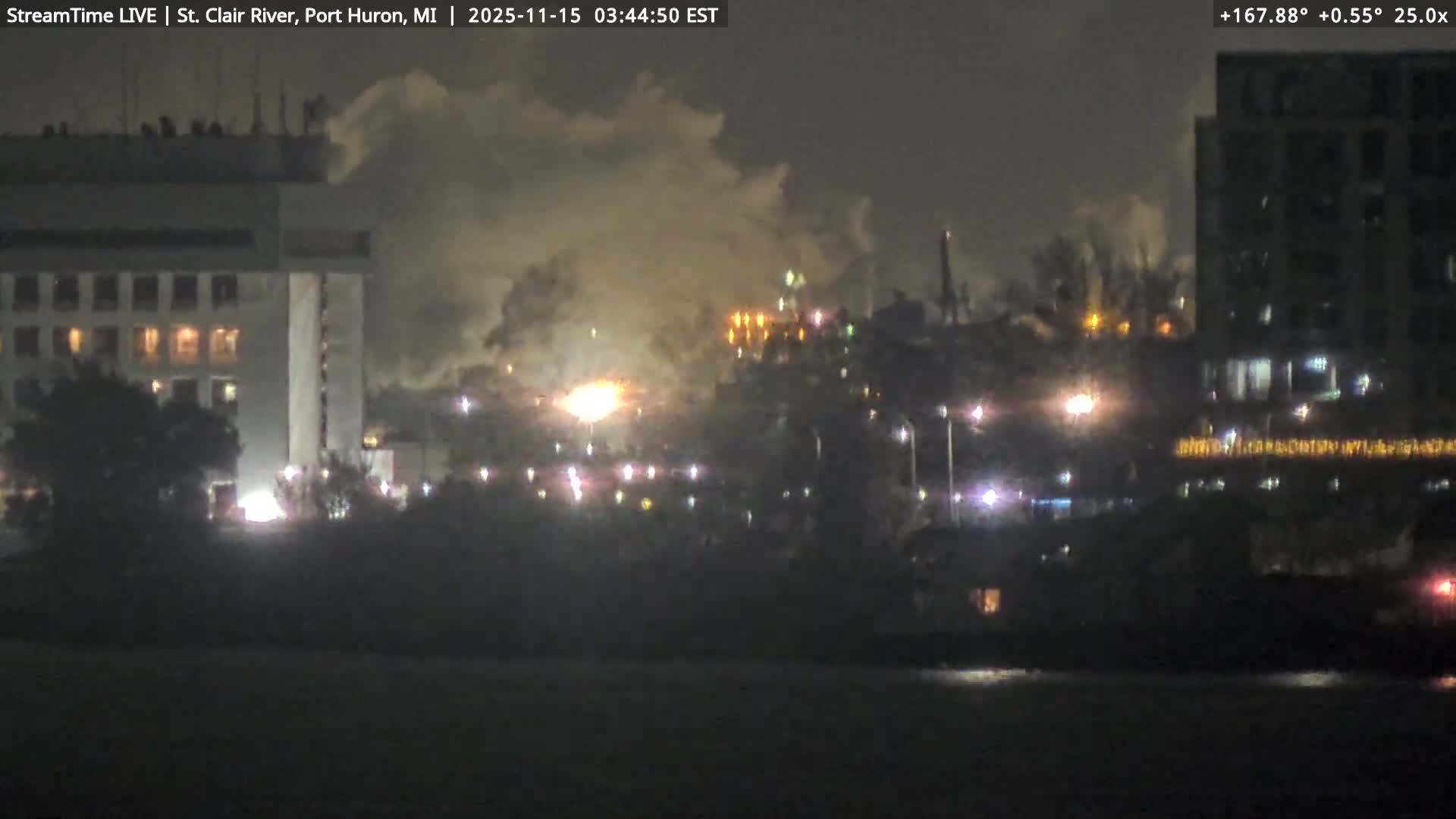 A hazy night view across a dark river shows a city's brightly lit buildings and an industrial area emitting large plumes of smoke or steam against an overcast sky.