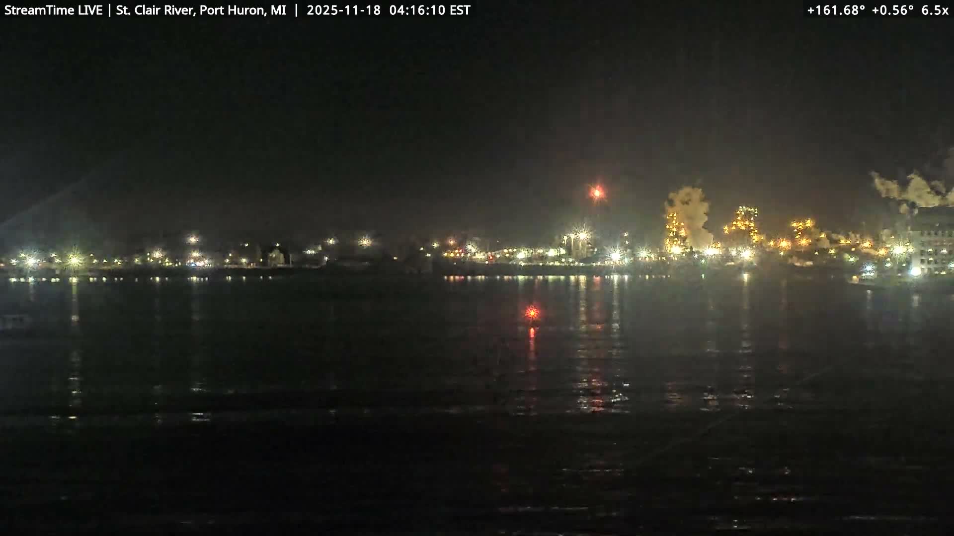 A hazy night view across a dark river shows a city's brightly lit buildings and an industrial area emitting large plumes of smoke or steam against an overcast sky.