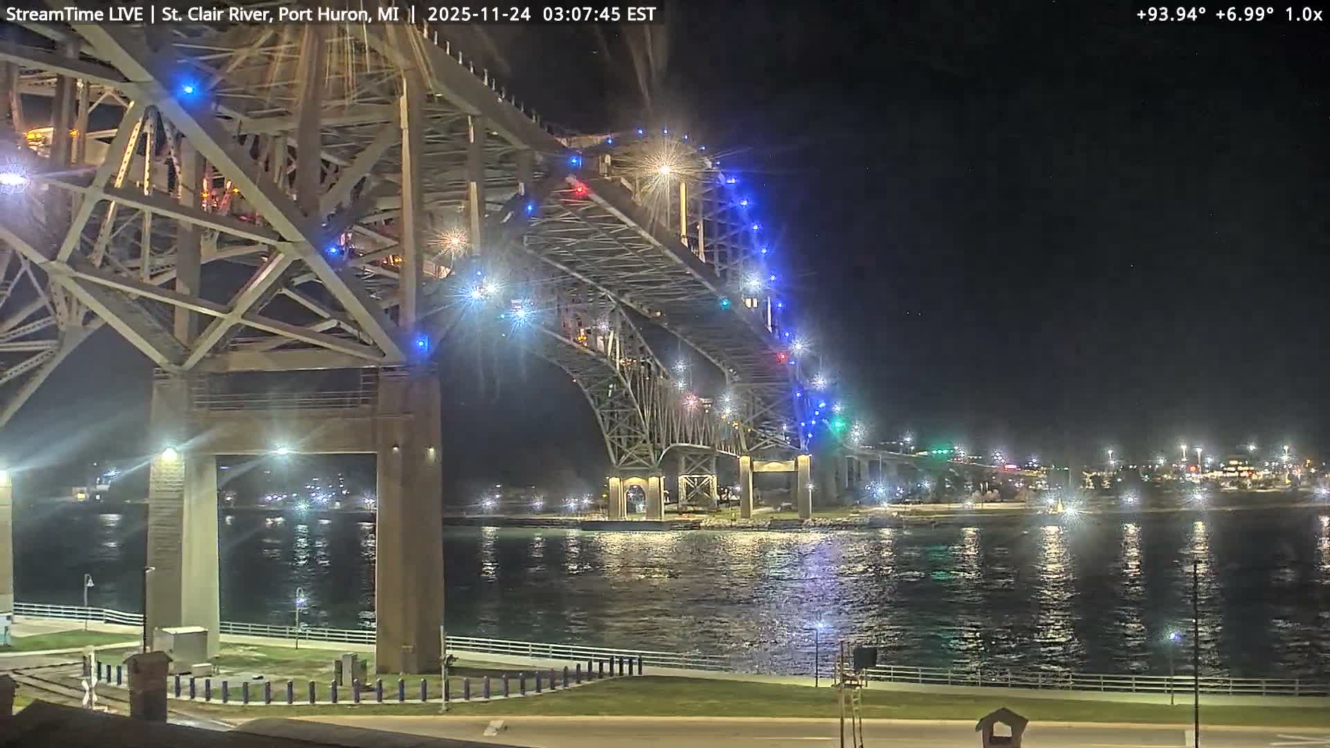 A large, brightly lit bridge adorned with colorful lights spans a calm body of water at night under a clear sky, with distant city lights visible on the far bank.