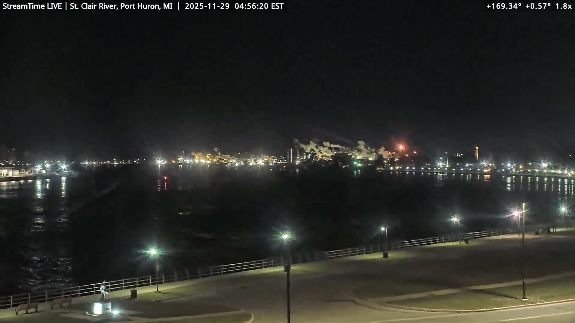 The image depicts a clear night over a river, with brightly lit industrial structures on the distant bank emitting plumes of smoke or steam, and a waterfront promenade with streetlights and a statue in the foreground reflecting lights on the water's surface.