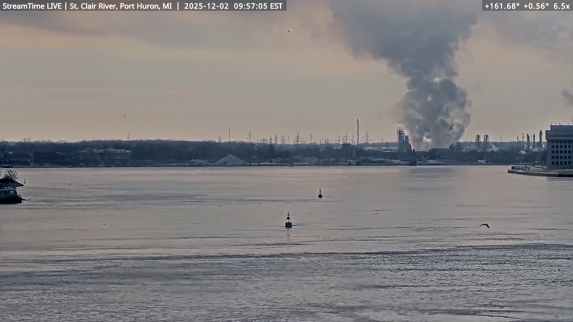 A wide, calm river with two buoys and several birds on the water stretches towards a distant industrial area on the far bank, which is emitting a large plume of dark smoke under an overcast, pale sky.