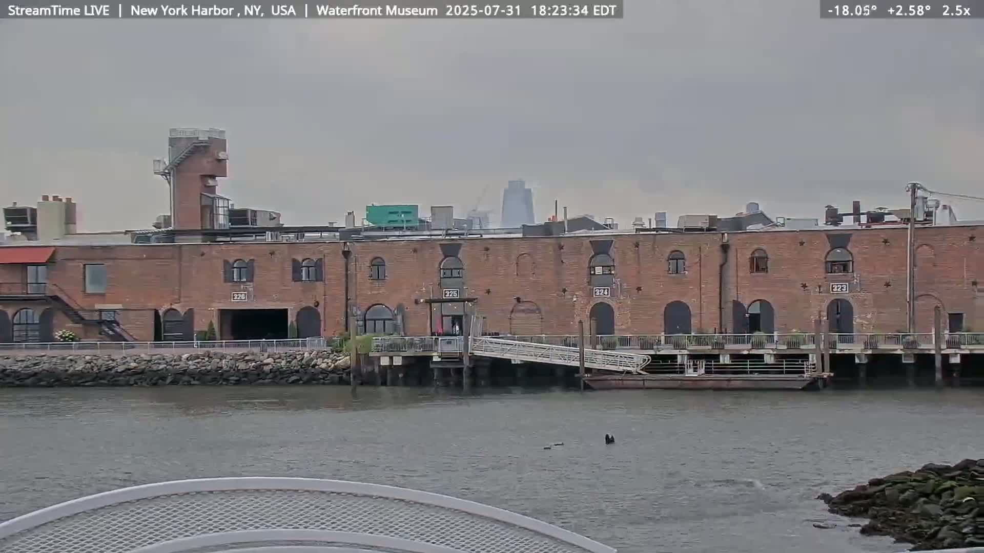 A long, brick building sits on the waterfront under an overcast sky, with a calm body of water in the foreground.