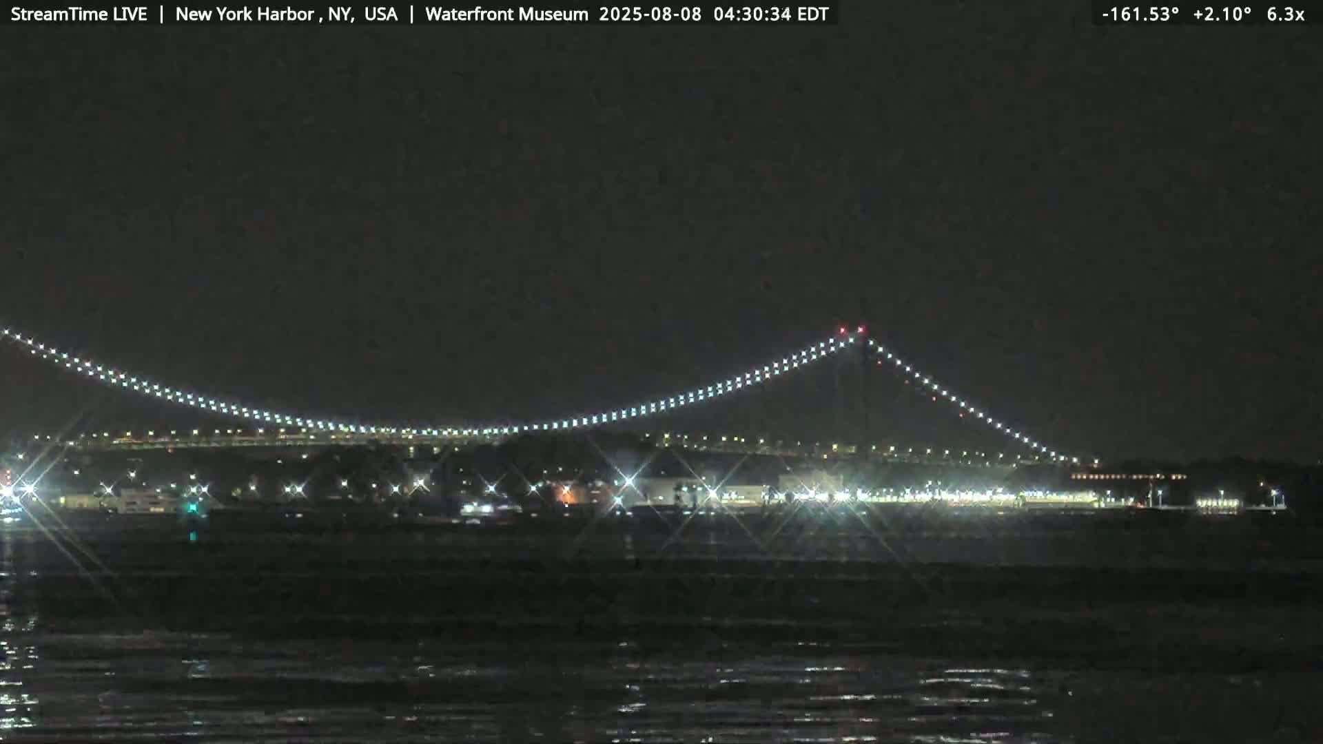 A nighttime view of a lit-up suspension bridge over dark water under a clear, dark sky.