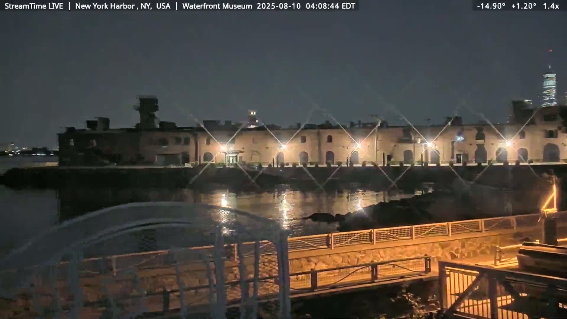 A nighttime view of a waterfront building complex illuminated by lights, reflecting on calm water, with a distant cityscape visible under a clear, dark sky.