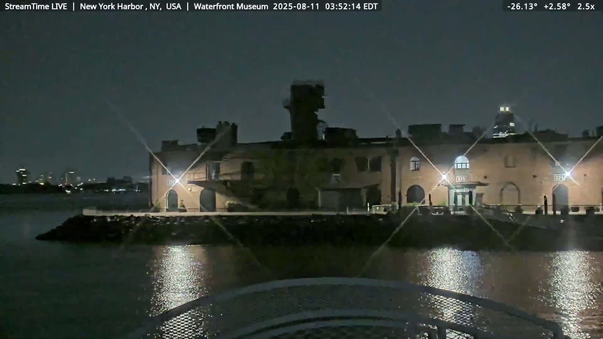A brick building, possibly a museum, sits on the water's edge at night, illuminated by lights, with calm water reflecting the lights.